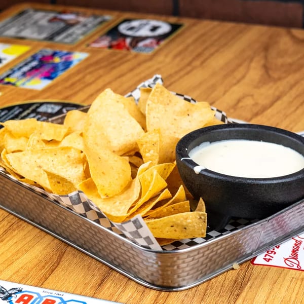 A metal tray filled with tortilla chips and a black bowl of white queso dip.