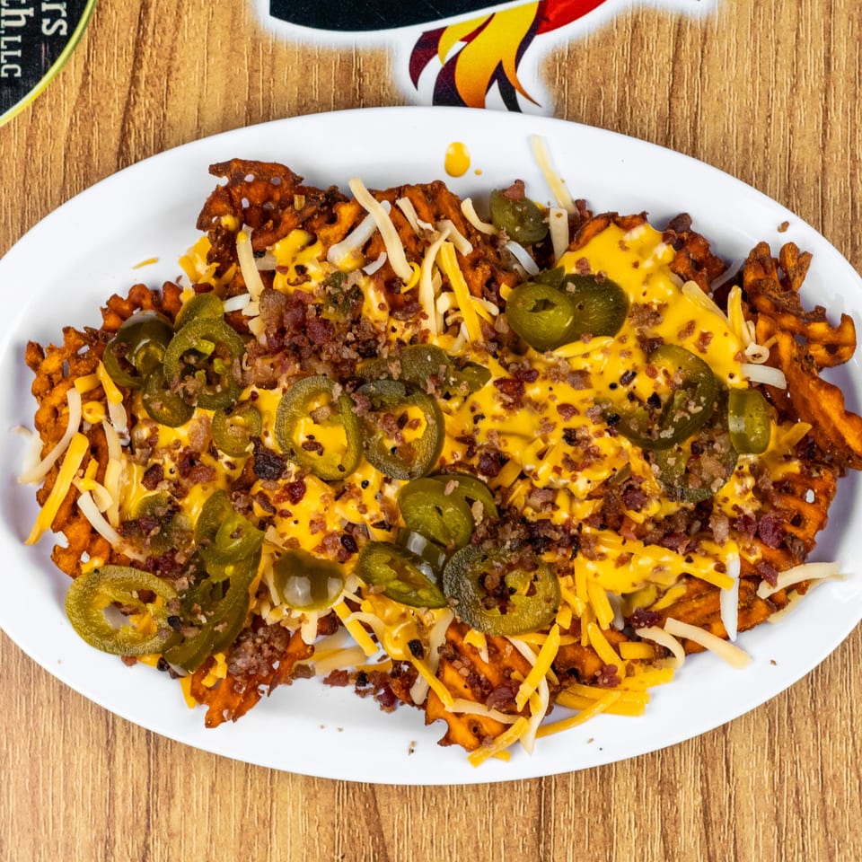 Waffle fries loaded with melted cheese, bacon bits, and sliced jalapeños on a white plate.