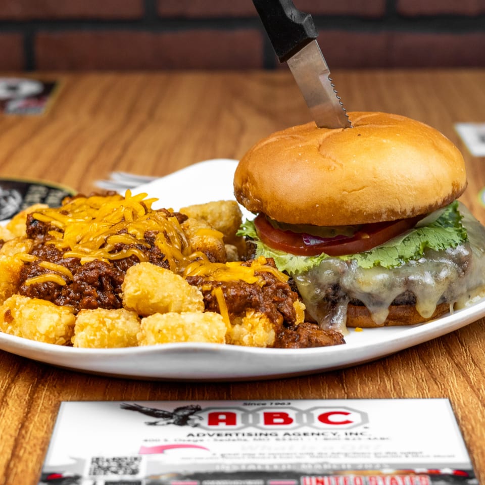 Cheeseburger with a knife, chili cheese tater tots, and a business card on a wooden table.